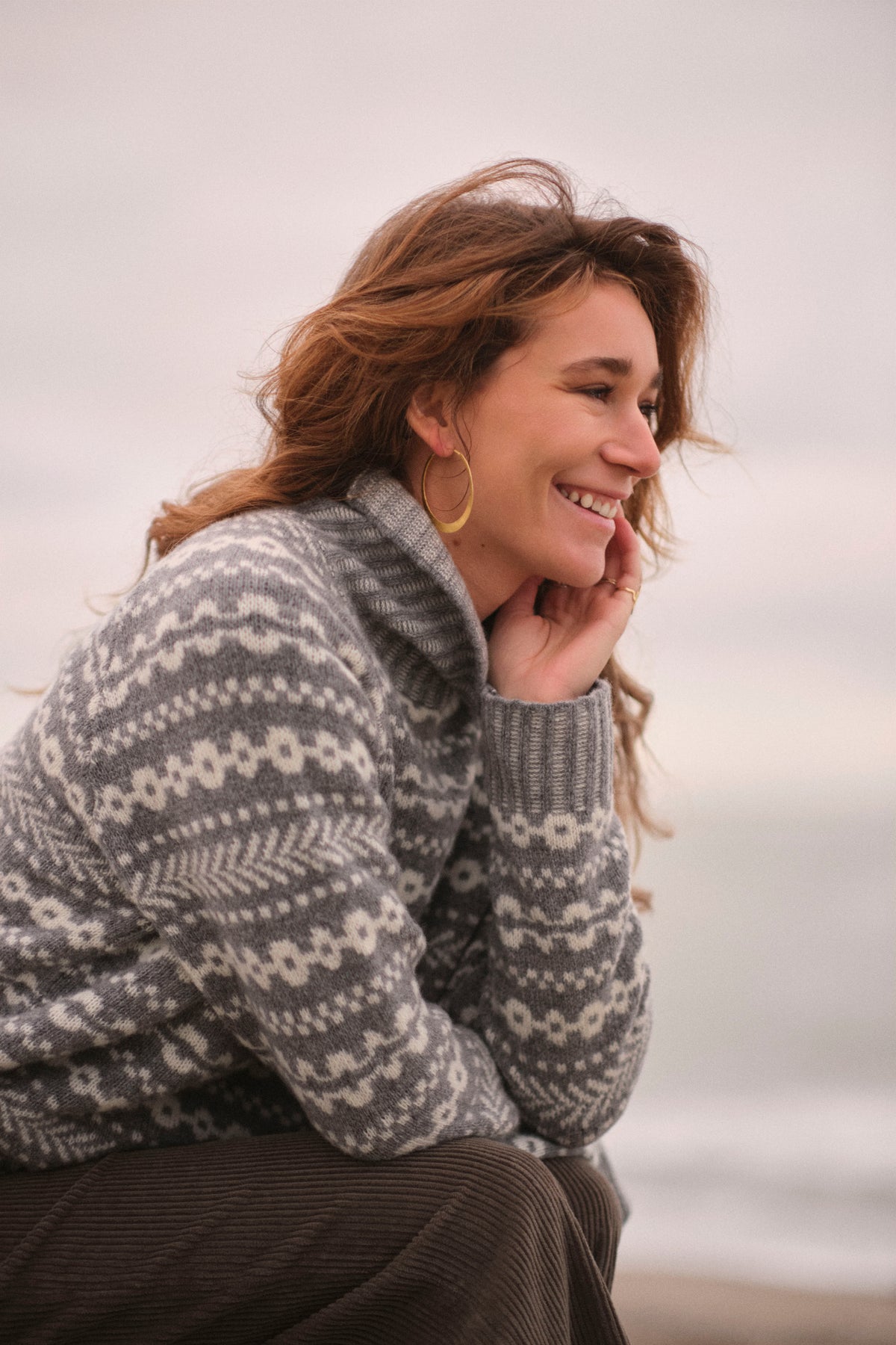 A woman with wavy light brown hair sits outdoors wearing a gray and white patterned knit sweater and dark brown corduroy pants, smiling and looking to the side against a soft, overcast sky.